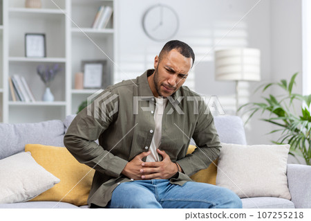 A young Muslim man grimaced in pain and holds his hands to the side of his stomach, suffering from appendicitis, convulsions. Sitting at home on the sofa, waiting for emergency medical help. A young Muslim man grimaced in pain and holds his hands to the side of his stomach, suffering from appendicitis, convulsions. Sitting at home on the sofa, waiting for emergency medical help. 107255218