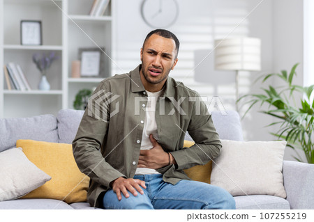 A young African American man is suffering from abdominal pain, stomach cramps and constipation. He is sitting on the sofa and holding his stomach, grimacing in pain. A young African American man is suffering from abdominal pain, stomach cramps and constipation. He is sitting on the sofa and holding his stomach, grimacing in pain. 107255219