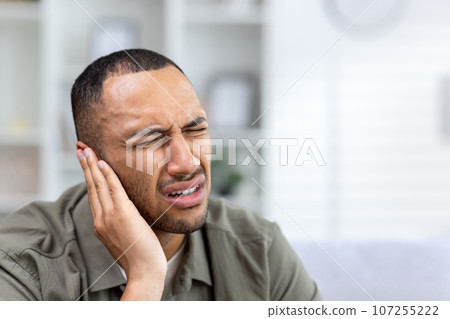 Unbearable and severe ear pain bothers a young African American man, sitting at home and holding his hand to his cheek, giving a massage. Close-up photo. Unbearable and severe ear pain bothers a young African American man, sitting at home and holding his hand to his cheek, giving a massage. Close-up photo. 107255222