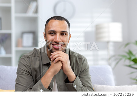 Portrait of a young African American man sitting on the couch at home with his hands folded together smiling at the camera. Close-up photo. Portrait of a young African American man sitting on the couch at home with his hands folded together smiling at the camera. Close-up photo. 107255229
