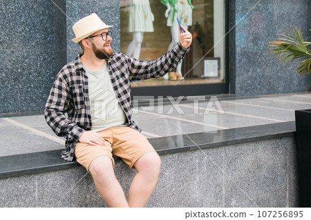 Bearded millennial man takes selfie via smartphone strolls in downtown poses against modern city building enjoys leisure time copy space. People urban lifestyle and technology concept 107256895