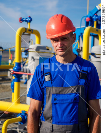Gas distribution station operator. Portrait of man in overalls and construction helmet on summer day Gas distribution station operator. Portrait of man in overalls and construction helmet on summer day 107259694