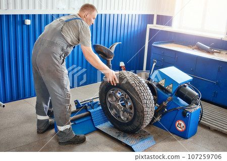 Mechanic works in tire repair shop. Car service mechanic puts wheel on car rim using special device Mechanic works in tire repair shop. Car service mechanic puts wheel on car rim using special device 107259706