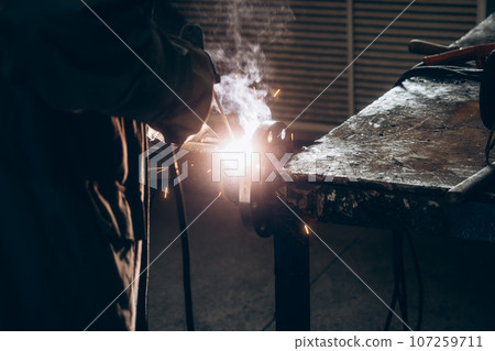 Welder works at workbench in welding shop. Flash. Electric arc. Dark photo. Background. Welder works at workbench in welding shop. Flash. Electric arc. Dark photo. Background. 107259711