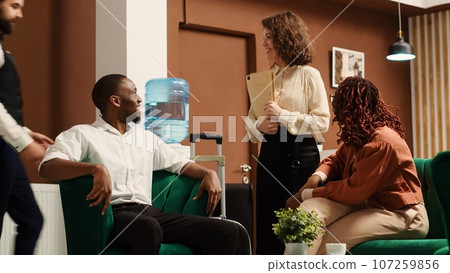 Positive friendly hotel manager greeting in tourists on vacation trip while they wait to be checked in their rooms. Relaxed guests happy with customer service provided by helpful concierge personnel Positive friendly hotel manager greeting in tourists on vacation trip while they wait to be checked in their rooms. Relaxed guests happy with customer service provided by helpful concierge personnel 107259856