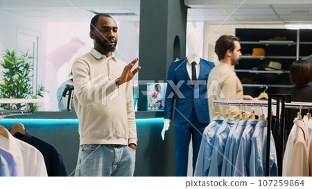 African american man looking at hologram in store, using augmented reality holographic image in fashion clothing boutique. Male customer checking clothes in shopping center. Tripod shot. 107259864