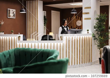 Hotel concierge writing booking form at front desk, preparing to help guests with accommodation at arrival. Female resort employee checking registrations in record, reception area. 107259889