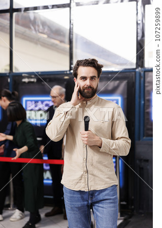 Professional TV news reporter standing with microphone against lined up shoppers. Journalist or correspondent preparing for live broadcast during Black Friday sales at local shopping mall 107259899