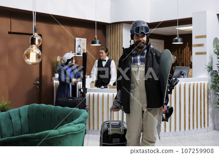 Male tourist wearing winter jacket and holding snowboard and suitcase leaves ski resort lounge area. Young man with snow gear heading to hotel room after check in while woman is assisted by staff. Male tourist wearing winter jacket and holding snowboard and suitcase leaves ski resort lounge area. Young man with snow gear heading to hotel room after check in while woman is assisted by staff. 107259908