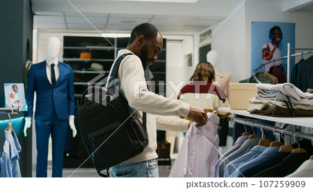 Male shopper buying new shirts for formal wear at mall, looking at new retail store merchandise to increase wardrobe. Young man checking trendy clothes in fashion boutique shop. 107259909