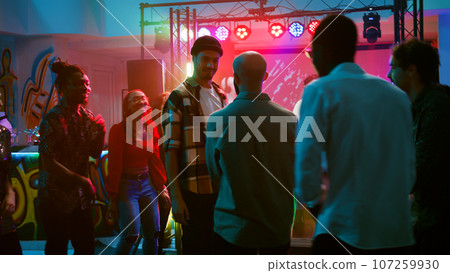 Happy adults improvise breakdance battle at club party, jumping around on dance floor with disco music and spotlights. Group of people showing off cool moves at nightclub. Handheld shot. Happy adults improvise breakdance battle at club party, jumping around on dance floor with disco music and spotlights. Group of people showing off cool moves at nightclub. Handheld shot. 107259930