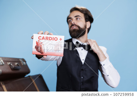 Hotel concierge presents cardiac pills in front of studio camera, recommending medication for heart disease or problems. Young man doorkeeper shows box of medicaments treatment. 107259958