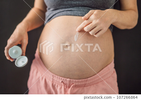 Positive young pregnant woman in comfortable homewear applying belly butter on her big tummy, woman belly 107266864