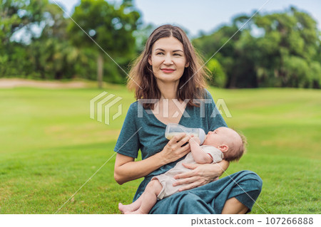 Mother holding and feeding baby from milk bottle in the park. Portrait of cute newborn baby being fed by her mother using bottle. Loving woman giving to drink milk to her boy 107266888