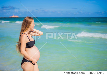 Radiant pregnant woman in a swimsuit, amid the stunning backdrop of a turquoise sea. Serene beauty of maternity by the shore Radiant pregnant woman in a swimsuit, amid the stunning backdrop of a turquoise sea. Serene beauty of maternity by the shore 107266942