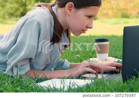 Teenage schoolgirl studying reading her books, tablet and notebook, sitting outdoors. Back to school. Student girl lying on the green grass using laptop in the college yard or park. Distance learning. Teenage schoolgirl studying reading her books, tablet and notebook, sitting outdoors. Back to school. Student girl lying on the green grass using laptop in the college yard or park. Distance learning. 107270397