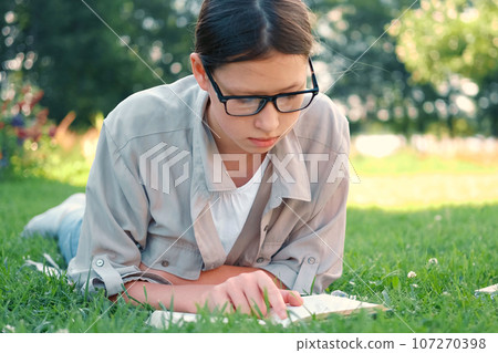 Teenage schoolgirl studying reading her books, lying down outdoors. Back to school, dreaming, inspiration. Student girl reads book on the green grass in the college yard or park. Distance learning. Teenage schoolgirl studying reading her books, lying down outdoors. Back to school, dreaming, inspiration. Student girl reads book on the green grass in the college yard or park. Distance learning. 107270398