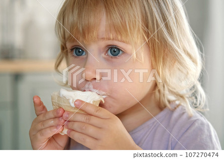 Baby girl enjoying ice cream. Pretty little toddler eating an ice-cream indoors, at home. Dining room background. Small child eats plombir and cream messy on her mouth. Cute kid with tasty sweet food. Baby girl enjoying ice cream. Pretty little toddler eating an ice-cream indoors, at home. Dining room background. Small child eats plombir and cream messy on her mouth. Cute kid with tasty sweet food. 107270474