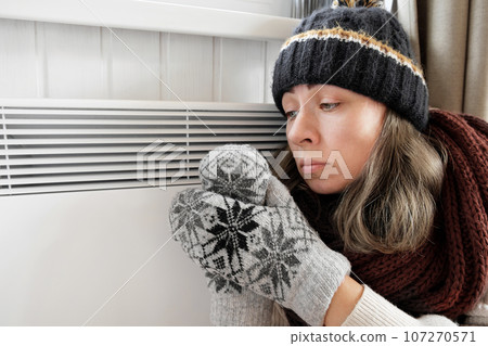 Young frozen woman wearing a sweater shaking and freezing for winter cold with shock expression on face. Girl is warming up hands with breath over electric heater. Discomfort spending time at home 107270571