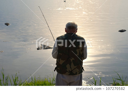 Old man fishing. Senior gray haired fisherman throws a spinning from shoreside at sunset, twisting a coil. Positive elderly male angling at lake, rotating reel rod. Retired fisher enjoying his weekend 107270582