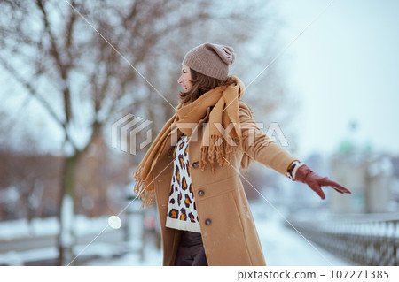 smiling stylish female in brown hat and scarf in camel coat 107271385