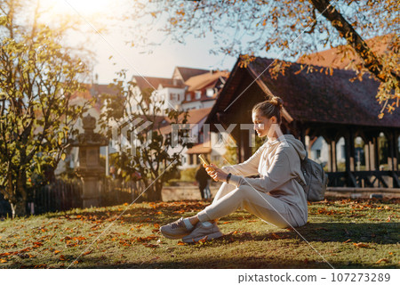 Young Fashionable Teenage Girl With Smartphone In Europian Park In Autumn Sitting At Smiling. Trendy Young Woman In Fall In Park Texting. Retouched, Vibrant Colors. Beautiful Blonde Teenage Girl 107273289