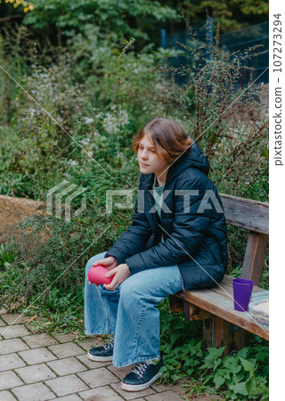 Blonde girl holding cup sitting on bench in the park. Autumn season. Young Teen Girl relaxing on bench with cup of coffee or tea. 107273294