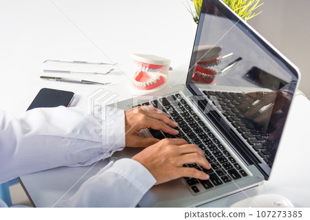 Dentist doctor or nurse in uniform use oral software teeth Xray and typing information medical history form into laptop computer for record document report, tooth model on desk, dentistry concept 107273385