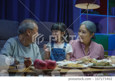 Happy Asian family grandmother grandfather and granddaughter dining on table and having fun during at home night time, senior parent and child eating food together in living room indoors together Happy Asian family grandmother grandfather and granddaughter dining on table and having fun during at home night time, senior parent and child eating food together in living room indoors together 107273402