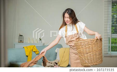 Beautiful young housekeeper maid doing housework holds basket wood of clean messy dirty clothes at home, Housewife woman holding basket with heap of different clothes on sofa, Laundry cleanliness Beautiful young housekeeper maid doing housework holds basket wood of clean messy dirty clothes at home, Housewife woman holding basket with heap of different clothes on sofa, Laundry cleanliness 107273535