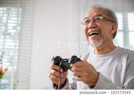 Asian senior old man enjoying holding joystick playing video game at home in living room, mature man hands using game controller, Funny on retirement elderly smile sitting on sofa life gaming 107273545