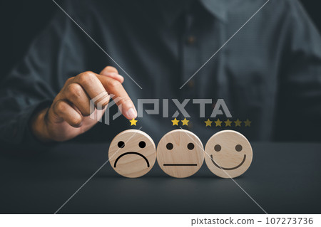 Negative customer experience concept. Hand holding wooden block with sad face icon on circle wooden table, symbolizing unhappiness and bad service. Dissatisfied client expressing dissatisfaction. Negative customer experience concept. Hand holding wooden block with sad face icon on circle wooden table, symbolizing unhappiness and bad service. Dissatisfied client expressing dissatisfaction. 107273736