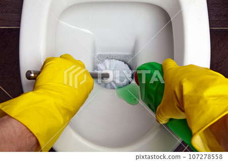 The hands of a person in yellow rubber gloves using a disinfectant gel and a toilet brush wash the toilet The hands of a person in yellow rubber gloves using a disinfectant gel and a toilet brush wash the toilet 107278558