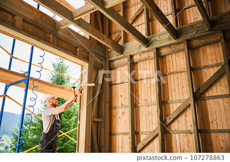 Carpenter constructing wooden frame, two-story house near forest. Bearded man hammering nails with hammer while wearing protective helmet and work coveralls. Concept of modern ecological construction. Carpenter constructing wooden frame, two-story house near forest. Bearded man hammering nails with hammer while wearing protective helmet and work coveralls. Concept of modern ecological construction. 107278863