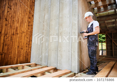 Carpenter constructing wooden framed house. Man worker cladding facade of house with cement particle boards, fastening them with screwdriver. Concept of modern eco-friendly construction. 107278866