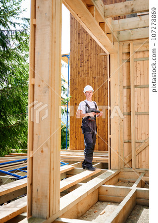 Portrait of carpenter constructing wooden framed house. Man worker cladding facade of house, using screwdriver. Concept of modern eco-friendly construction. Portrait of carpenter constructing wooden framed house. Man worker cladding facade of house, using screwdriver. Concept of modern eco-friendly construction. 107278869