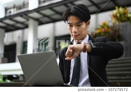 Young businessman using laptop and checking time on wristwatch for his next appointment 107279572