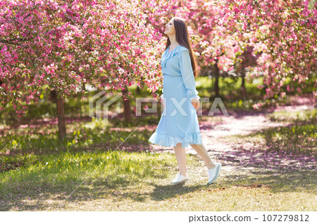 Happy girl in blue dress, is standing in a pink blooming garden 107279812