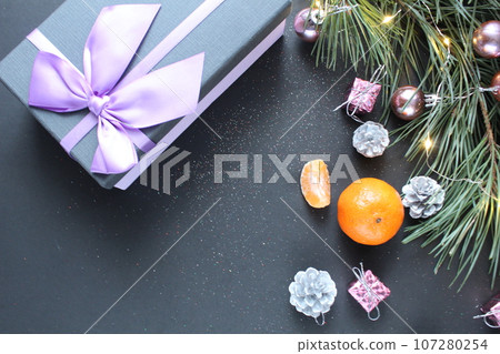 still life New Year's Christmas. A gift with a bow lies on a black background next to a branch of spruce pine trees mandarin toys Christmas tree with a place for text copyspace top view still life New Year's Christmas. A gift with a bow lies on a black background next to a branch of spruce pine trees mandarin toys Christmas tree with a place for text copyspace top view 107280254