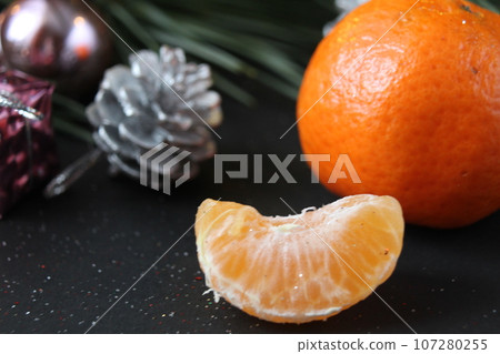mandarin whole in the peel and a slice of fruit lie side by side on a black background next to a branch of spruce and a silver cone. New Year and Christmas. holiday attribute mandarin whole in the peel and a slice of fruit lie side by side on a black background next to a branch of spruce and a silver cone. New Year and Christmas. holiday attribute 107280255
