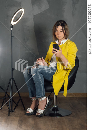 Concentrated lady in yellow coat looking at screen of phone sitting on chair near annular LED lamp against gray wall. Concentrated lady in yellow coat looking at screen of phone sitting on chair near annular LED lamp against gray wall. 107280928