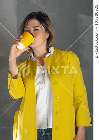 Charming lady in yellow coat drinking hot beverage from xanthous paper coffee cup standing against gray background. Charming lady in yellow coat drinking hot beverage from xanthous paper coffee cup standing against gray background. 107280929
