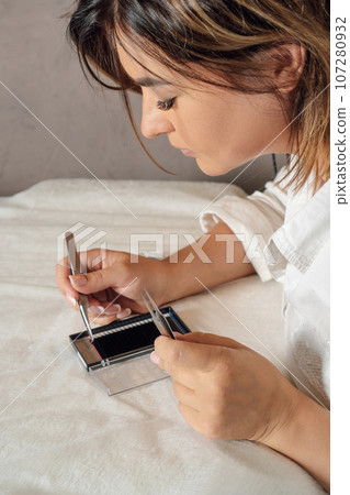 Gorgeous lashmaker looking at palette of black artificial eyelashes and picking material for procedure with tweezers. Gorgeous lashmaker looking at palette of black artificial eyelashes and picking material for procedure with tweezers. 107280932