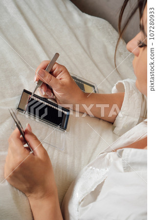 Focused lashmaker with tweezers in hands carefully taking black artificial eyelash from pallet lying on white blanket. Focused lashmaker with tweezers in hands carefully taking black artificial eyelash from pallet lying on white blanket. 107280933