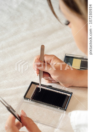 Close up of concentrated lashmaker taking black artificial eyelash from palette using tweezers that holding in hands. Close up of concentrated lashmaker taking black artificial eyelash from palette using tweezers that holding in hands. 107280934