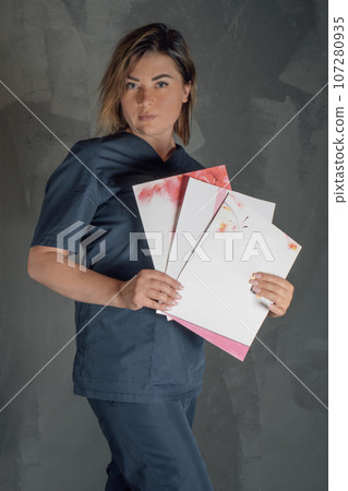 Young female professional holding three certificates in hand and pointing to documents standing against gray wall. Young female professional holding three certificates in hand and pointing to documents standing against gray wall. 107280935