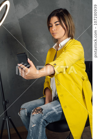 Stylish young lady looking at camera, taking selfie sitting on chair next to annular LED lamp against gray background. Stylish young lady looking at camera, taking selfie sitting on chair next to annular LED lamp against gray background. 107280939
