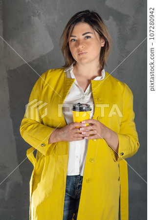 Delightful young woman in bright yellow coat standing against gray wall indoors with xanthous coffee cup in hands. 107280942