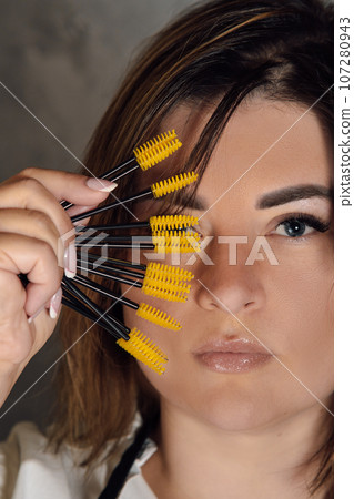 Gorgeous young lashmaker holding yellow disposable eyelash extension brushes close to face standing on gray background. 107280943