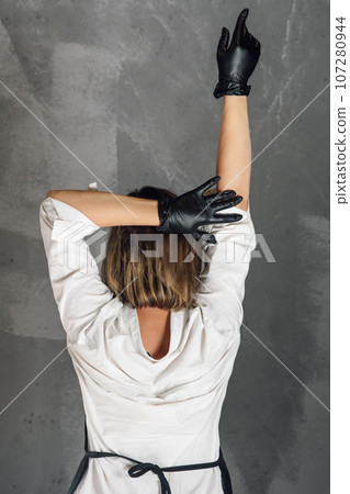 Rear view of lady in black medical gloves standing against gray wall, lifting one arm, holding other hand behind head. Rear view of lady in black medical gloves standing against gray wall, lifting one arm, holding other hand behind head. 107280944